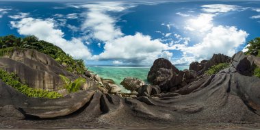 Yükselen granit kayalar, güneşli bir gökyüzünün altındaki engebeli kıyı şeridini saran yemyeşil yemyeşil sularla denizin durgun turkuaz sularına açılır. Anse Kaynak dArgent. La Digue, Seyşeller. VR 360.