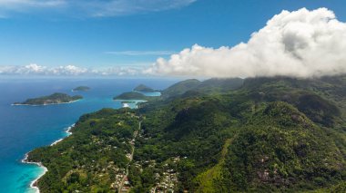 Yeşil yemyeşil tepeler bulutlu bir gökyüzüyle turkuaz sulara doğru uzanıyor. Seyşeller, Mahe.