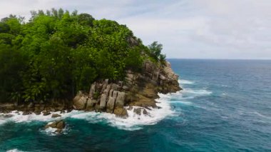 Yoğun yeşil bitki örtüsü sarp bir yamacı kaplıyor. Bu yamaç turkuaz okyanusla birleşiyor ve dalgalar kayalara çarpıyor. Seyşeller, Mahe.