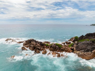 Yoğun yeşil yapraklar ve uzak tepelerle çevrili büyük kayalık oluşumların turkuaz sularla buluştuğu bir kıyı manzarası. Praslin, Seyşeller.