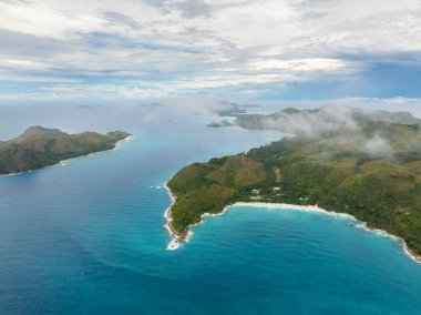 Ufuk boyunca uzanan turkuaz suları ve kayalık bir kıyı şeridi ile sisli, yemyeşil tepelerin havadan görünüşü. Praslin, Seyşeller.