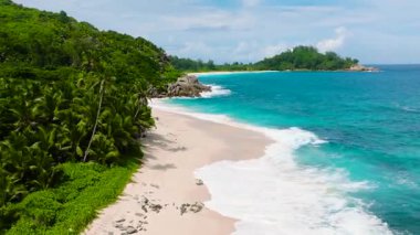 Granit kayalar tropikal bir kıyıda duruyor, dalgalar onlara çarpıyor ve arka planda yoğun yeşil yapraklar var. Seyşeller, Mahe. Anse Boileau..