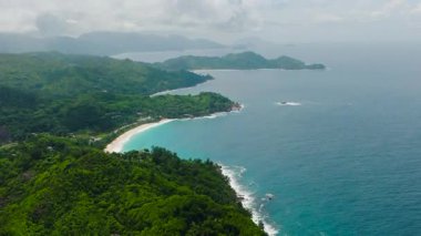 Yeşil bitkilerle kaplı tepeler, kıyı boyunca parçalı bulutlu bir gökyüzünün altında turkuaz okyanusla buluşuyor. Seyşeller, Mahe.