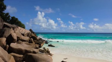 Dalgalar açık bir gökyüzünün altında kumlu sahile yavaşça yaklaşırken granit kayalar okyanusa doğru uzanıyor. Büyük Anse. Seyşeller, La Digue.