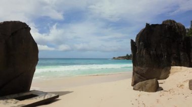 Turkuaz okyanus dalgaları kumlu bir sahilde kayalara çarpıyor ve arka planda yemyeşil bir alan var. Anse Niyet. Seyşeller, Mahe.