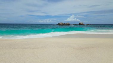 Dalgalar yavaşça kumlu kıyıya doğru yuvarlanırken büyük kayalar parlak mavi bir okyanusa karşı görülebilir. Seyşeller, Mahe. Anse Boileau.