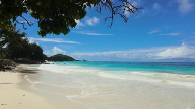 Turkuaz su, kumlu sahille yemyeşil ve mavi gökyüzünün altındaki uzak tepelerle buluşur. Seyşeller, Praslin.