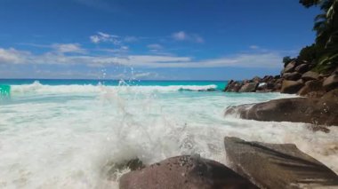 Dalgalar açık mavi gökyüzünün altında turkuaz sularla tropikal bir sahilde büyük granit kayalara çarpıyor. Seyşeller, Praslin. Anse Georgette.