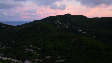 Dağılmış evleri ve pembe bir gökyüzünün altında kıyı manzarası olan bereketli bir dağ manzarası. Seyşeller, Mahe.