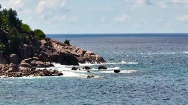 Parçalı bulutlu bir gökyüzünün altında yemyeşil ve köpüklü okyanus dalgalarının yer aldığı Rocky kıyı şeridi. Seyşeller, Mahe.