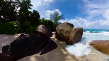 Okyanus dalgalarının parlak bir gökyüzünün altında dalgalandığı kumlu bir sahil boyunca devasa kayalar. Seyşeller, Mahe. Anse Takamaka.