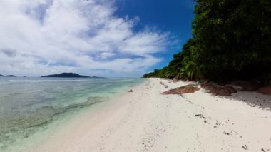 Yoğun yeşil bitki örtüsü, turkuaz okyanus dalgaları ve uzak adaları olan açık mavi bir gökyüzü ile çevrili beyaz kumlu sahil. Seyşeller, La Digue.