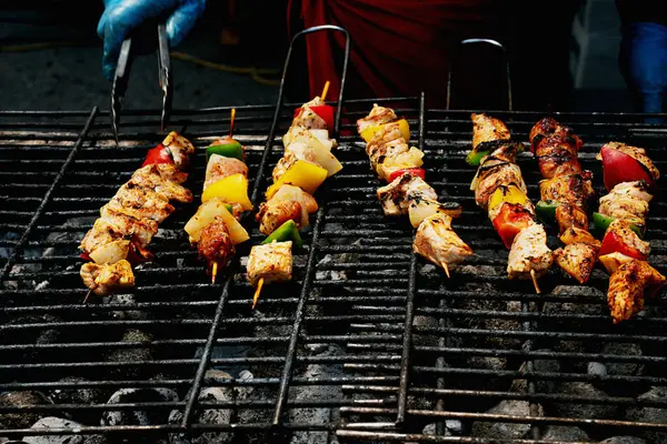 stock image Person in gloves turning skewers of marinated meat and vegetables on a hot grill, with smoke rising.