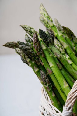 Hasır bir sepette, tarafsız bir arkaplanda duran taze kuşkonmaz demetlerinin yakın plan fotoğrafları. Kuşkonmaz mızrakları mor uçlu canlı yeşildirler. Tazeliklerini ve kalitelerini gösterirler..