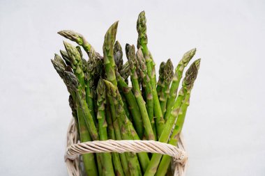 Hasır bir sepette, tarafsız bir arkaplanda duran taze kuşkonmaz demetlerinin yakın plan fotoğrafları. Kuşkonmaz mızrakları mor uçlu canlı yeşildirler. Tazeliklerini ve kalitelerini gösterirler..