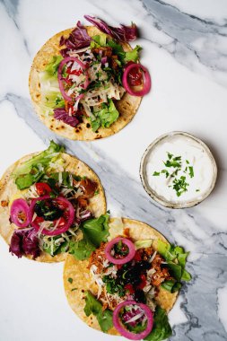 Karışık yeşillikli üç taco, soğan turşusu ve mermer bir yüzeyde servis edilen dilimlenmiş peynir..