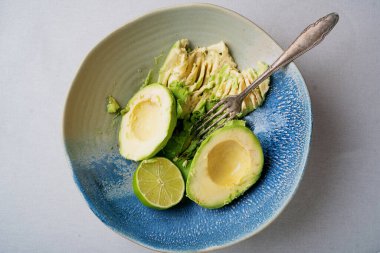 Üzerinde yarım avokado, yarım limon ve yanında biraz avokado püresi olan klasik çatal bulunan mavi ve bej bir seramik kase. Açık renkli tekstil yüzeyi. Üst Görünüm