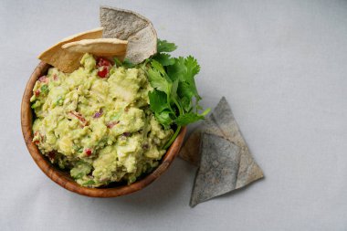 Kişniş sosuyla süslenmiş ve arka planda cipslerle servis edilen taze yapılmış guacamole kasesi. Guacamole, domates, soğan ve avokado parçalarıyla doludur..