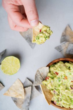 Guacamole dolu ahşap bir kasenin üzerinde kişniş soslu tortilla cipsi tutuyorum. Limon dilimi ve mavi mısır cipsi etrafa saçılmış..