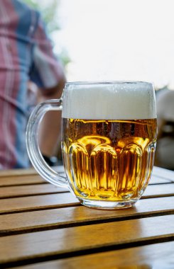  Elinde köpüklü, ahşap bir masada duran cam bardak dolusu altın bira. Soğuk bir içeceğin ferahlatıcı esansını arka planda bulanık bir şekilde açık havada yakalıyorum..