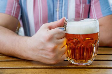  Elinde köpüklü, ahşap bir masada duran cam bardak dolusu altın bira. Soğuk bir içeceğin ferahlatıcı esansını arka planda bulanık bir şekilde açık havada yakalıyorum..