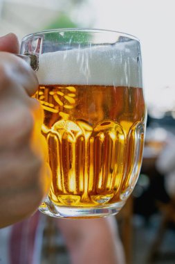  Elinde köpüklü, ahşap bir masada duran cam bardak dolusu altın bira. Soğuk bir içeceğin ferahlatıcı esansını arka planda bulanık bir şekilde açık havada yakalıyorum..