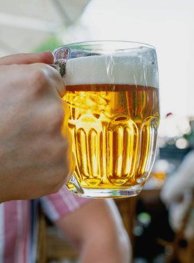  Elinde köpüklü, ahşap bir masada duran cam bardak dolusu altın bira. Soğuk bir içeceğin ferahlatıcı esansını arka planda bulanık bir şekilde açık havada yakalıyorum..