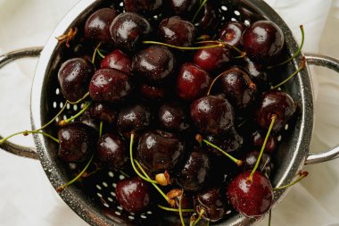 Yukarıdan yakalanmış, metalik bir kevgirde taze kirazlar. Su damlacıkları canlı görünümlerini güçlendirir. Üst görünüm beyaz kumaş arkaplanındaki parlak meyveyi vurgular.