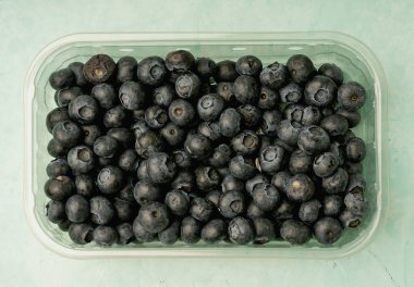 Açık mavi arka planda, şeffaf plastik bir kabın içindeki taze yaban mersinlerinin üst görüntüsü. Koyu mavi tonlar meyvenin doğal dokusunu ve olgunluğunu vurgular.