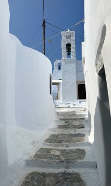 Ege Denizi 'ndeki bir Yunan adası olan Amorgos' ta mavi kapılı, taş basamaklı ve çan kuleli dar sokaklar. Parlak gökyüzü, geleneksel Kiklad mimarisi ve sakin atmosfer.