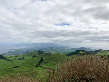 Azores Adası Sao Miguel manzarası. Yuvarlanan yeşil tepeler, yoğun ormanlar, bulutlu gökyüzünün altında volkanik göl manzarası. Doğayla, dağlarla ve canlı bitkilerle büyüleyici manzara ufku.