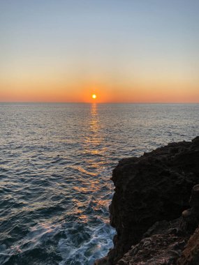Sakin okyanus sularının üzerinde gün batımında altın yansımalar ve ön planda kayalık kayalıklar. Güneş ufukta parlıyor ve sakin bir deniz manzarası yaratıyor.