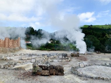 Yeşil tepeler, buhar kolonları, kayalık zemin ve arka planda tahta bir yapı ile çevrili çorak bir alandaki jeotermal bacalardan yükselen volkanik buhar Azores Adası Sao Miguel.