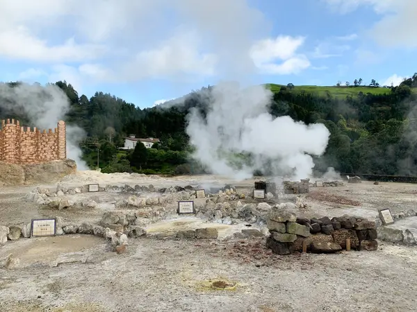 Yeşil tepeler, buhar kolonları, kayalık zemin ve arka planda tahta bir yapı ile çevrili çorak bir alandaki jeotermal bacalardan yükselen volkanik buhar Azores Adası Sao Miguel.