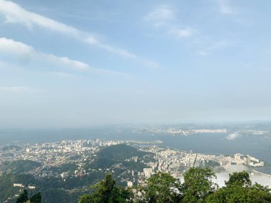 Rio de Janeiro şehir manzarasının tepesinde dağlar, körfez ve parlak gökyüzü var. Manzaralı hava perspektifi şehir manzarasını ve doğal çevreyi gösteriyor.