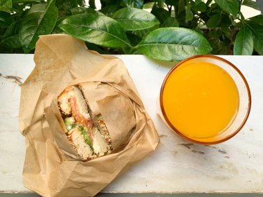 Kahverengi kağıda sarılı taze simit sandviçi ve bir bardak portakal suyu. Arka planda doğal yeşillik olan parlak kompozisyon, kahvaltı ya da atıştırmalık kavramları için mükemmel..