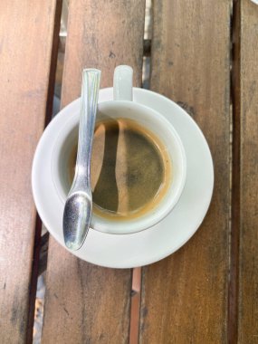 Beyaz fincanda sade bir kahvenin en üst görüntüsü. Kırsal ahşap bir masada kaşıkla doğal ve rahat bir açık hava ortamı yaratır. Basit ama davetkar kompozisyon.