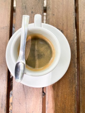 Beyaz fincanda sade bir kahvenin en üst görüntüsü. Kırsal ahşap bir masada kaşıkla doğal ve rahat bir açık hava ortamı yaratır. Basit ama davetkar kompozisyon.