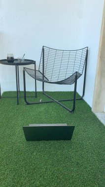 Siyah bir sandalyesi, küçük bir masası ve yeşil suni çimlerin üzerinde beyaz bir duvara dayalı laptopu olan minimalist çalışma alanı. Modern tasarım ve doğal ışık.