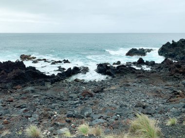 Sao Miguel 'in engebeli kıyı şeridi, Azores, volkanik kayalar ve çarpan dalgalarla. Uçurumların ve bitki örtüsünün ortasındaki okyanusa giden bir yürüyüş yolu, deniz kenarında doğal bir manzara sergiliyor..
