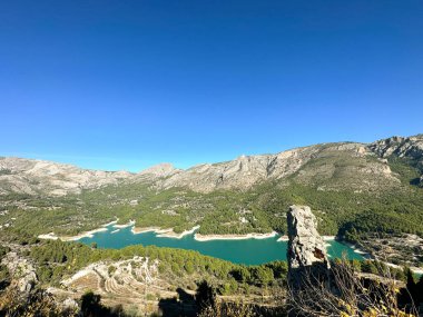 Turkuaz göl kenarındaki Guadalest manzarası açık mavi gökyüzünün altında kayalık dağlar ve yoğun yeşil ormanlarla çevrili. İkinci fotoğrafın ön planında geleneksel terracotta çatıları.