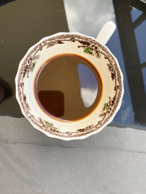 Yansıtıcı siyah bir yüzey üzerinde kahveyle dolu klasik bir seramik bardağın üst görüntüsü. Karmaşık çiçek deseni, bardağın kenarını gökyüzü yansımasıyla süslüyor..