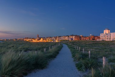 Katwijk aan Zee, Hollanda - 11 Haziran 2022: Kum tepelerinden bulvara yürüme yolu olan Katwijk aan Zee 'de mavi saatle gün batımı.