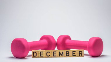 Ahşap bloklar beyaz arka planda pembe bir dambılın önünde 'Aralık' ı heceliyorlar. Bu minimalist kompozisyon fitness hedeflerini, motivasyonu ve yeni bir başlangıcı simgeliyor.