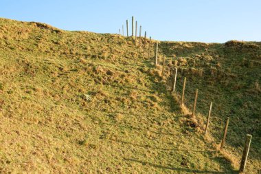 Fence leading up and over slope under blue sky in agricultural background.