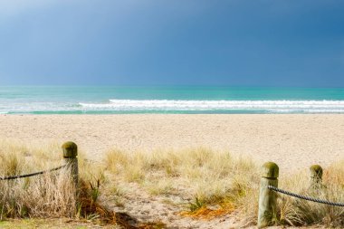 Sun illuminates sand beach under dark cloud with white water of breaking waves at Mount Maunganui in tauranga New Zealand.