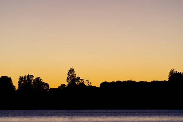 Land and trees in silhouette on horizon at sunrise across bay.