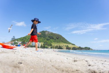Tauranga New Zealand- December 21 2015; Boy in blue sun hat and red shorts walks down Mount Maunganui beach clipart