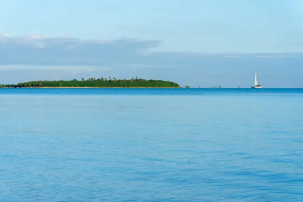 Distant small yacht under sail way off on horizon sailing toward small island in Pacific ocean near Fiji.