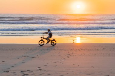 Sörfçüler Cenneti - 21 Eylül 2023; Sörfçüler Paradise plajında uzak sularda önemsiz insanlarla gün doğumu. Ve kum üzerinde sürerken hareket eden bulanık bisikletçi..